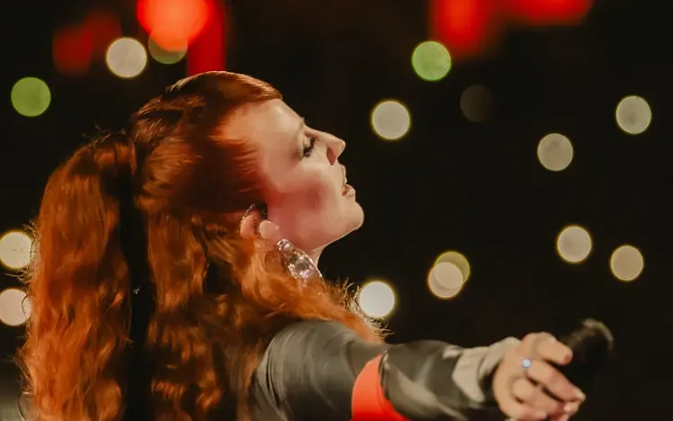 Jess Glynne in front of sparkly light with a mic in her hand